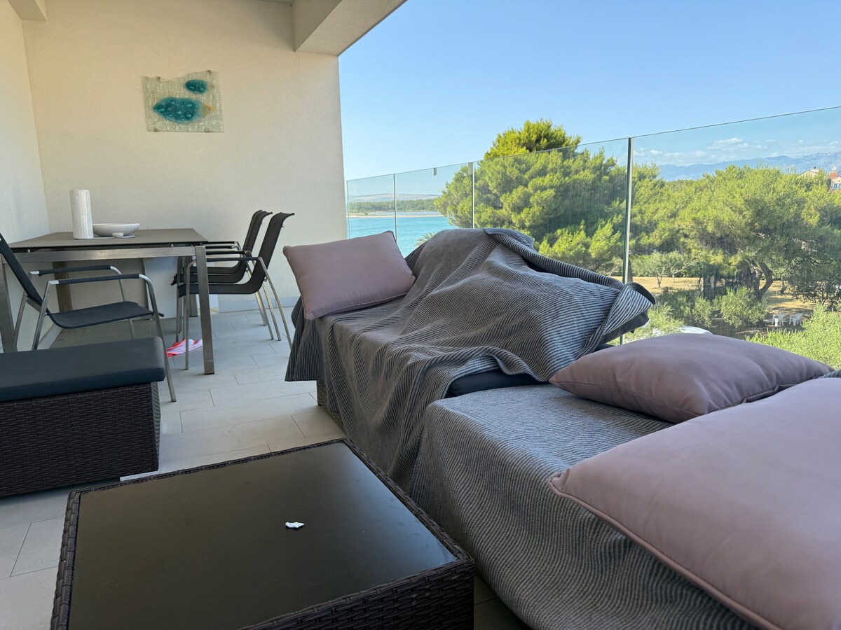 A covered balcony offers a view of the water and greenery. Comfortable seating is arranged with plush cushions and a blanket draped over a lounge chair. A modern dining table is set nearby, complemented by minimalist black chairs.