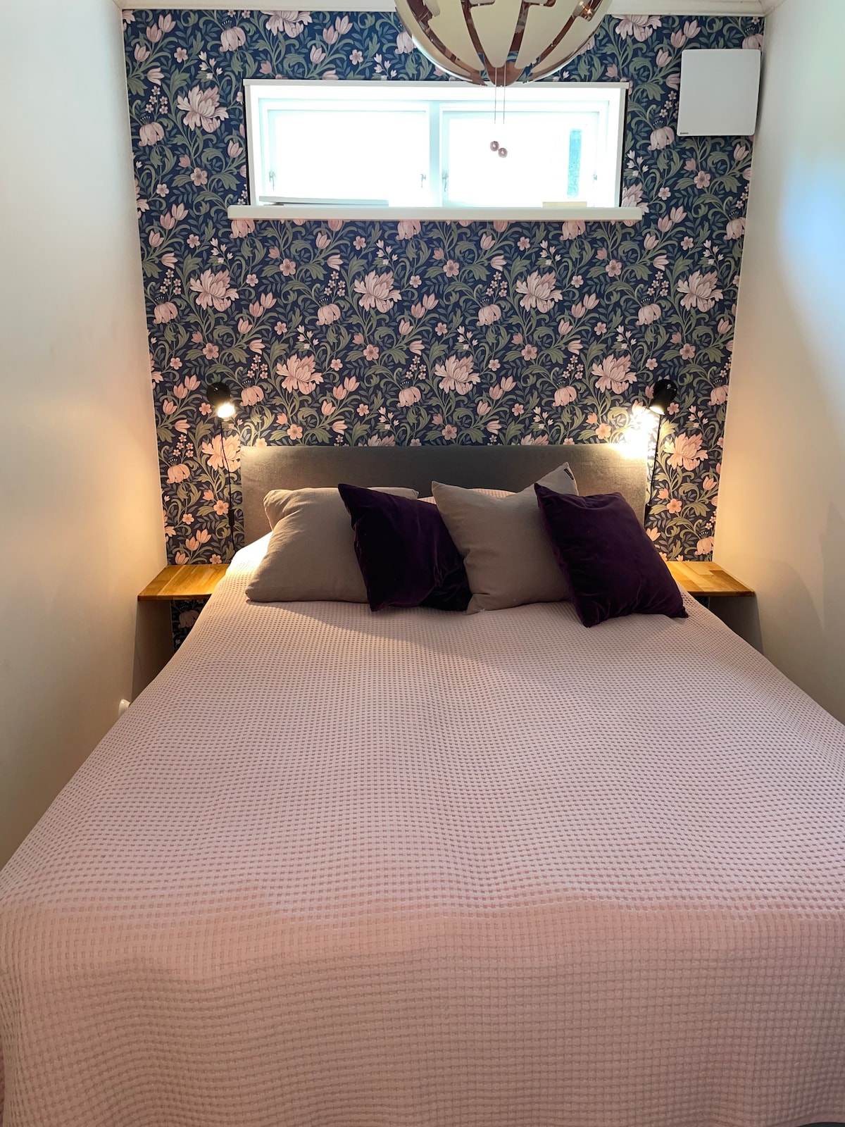 في غرفة النوم الصغيرة، يوجد سرير بعرض 140 سم مع طاولات بجانب السرير ومصابيح للقراءة 