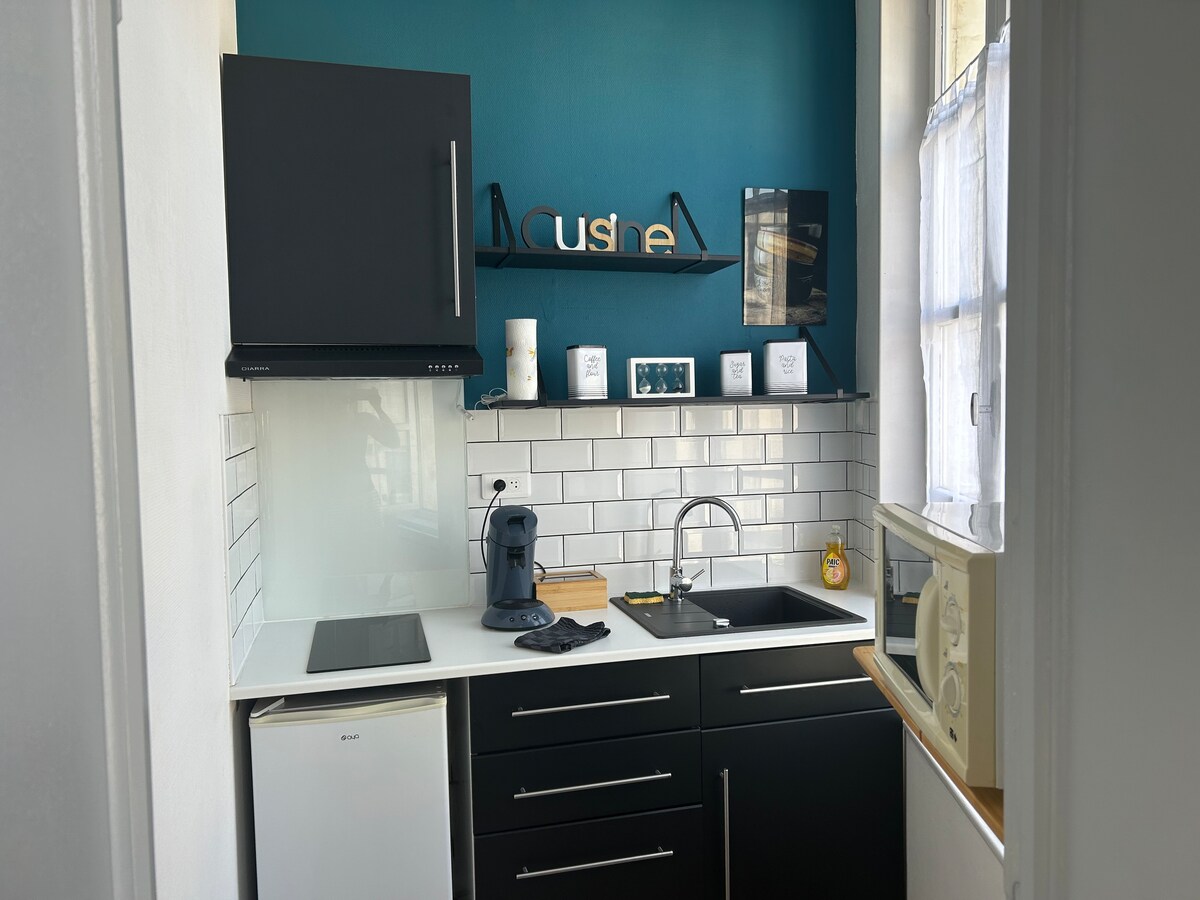 The compact kitchen features modern black cabinetry and a contrasting teal accent wall. A sleek sink is positioned beside a microwave and a small refrigerator. Counter space includes a coffee machine and a decorative shelf with framed pictures and essential kitchen items.