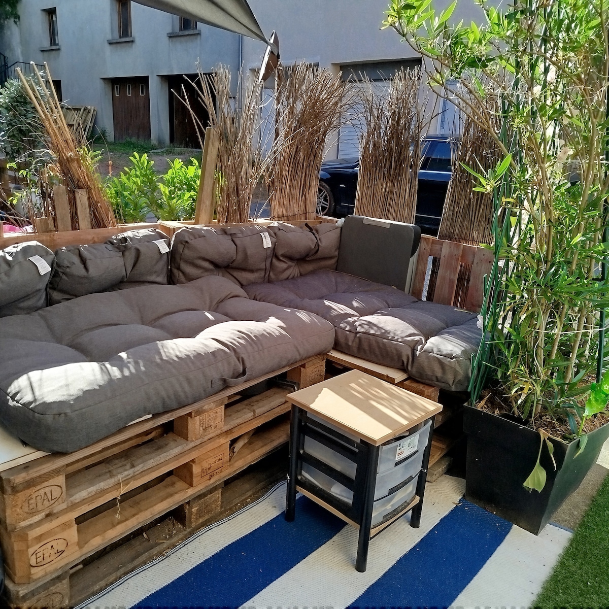 An outdoor seating area is composed of cushioned seating arranged on wooden pallets, providing a relaxed space. A small side table stands nearby, accompanied by a pot with lush green plants. Natural elements enhance the inviting atmosphere of this gathering spot.