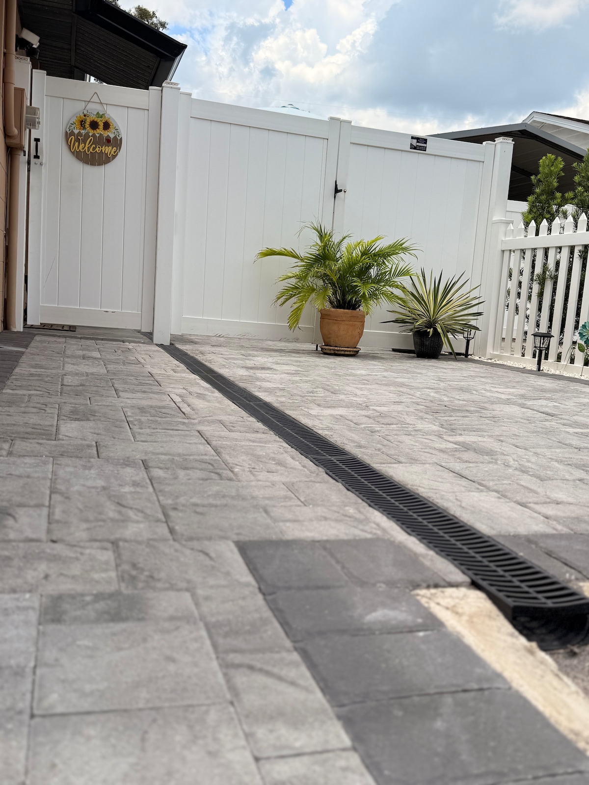 An outdoor area features a paved surface with a blend of grey and black tiles. A welcoming sign hangs on a white fence, which encloses the space. Potted plants add greenery, including a tall palm and another succulent, enhancing the relaxing environment.
