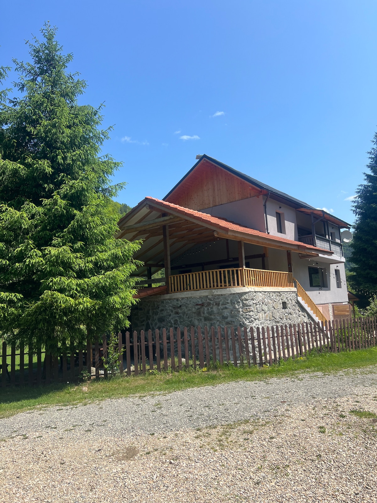 A two-story cottage is nestled among tall evergreen trees, featuring a stone and wooden facade. A covered balcony extends from the upper level, offering ample outdoor space. A gravel path leads to the welcoming entrance, framed by a wooden fence surrounding the property.