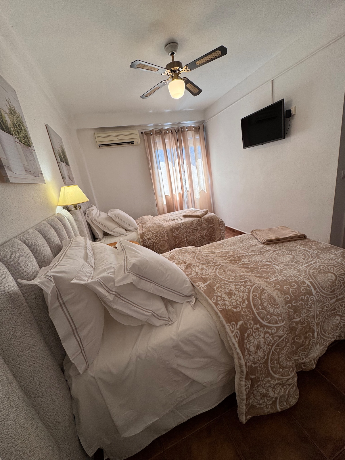 A serene bedroom features two spacious beds adorned with neutral-toned bedding. A ceiling fan provides gentle airflow, while a flat-screen TV is mounted on the wall. Natural light filters through sheer curtains, enhancing the overall brightness of the room.