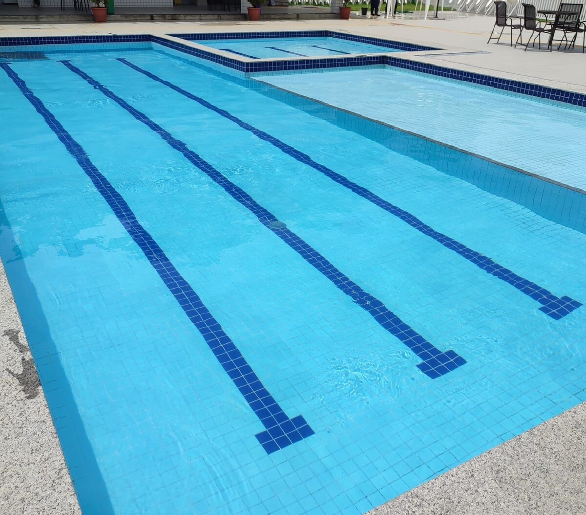A serene swimming area features a large pool with clearly marked lanes, showcasing pristine blue tiles reflecting sunlight. A smaller adjoining pool is visible nearby, enclosed by a patio area with scattered seating arrangements.