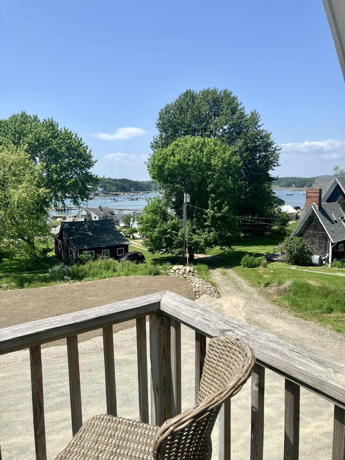 A balcony offers seating for two, overlooking a scenic view of Bass Harbor and the surrounding greenery. Large trees shade the area, and glimpses of water are visible among the landscape. A calm atmosphere is created by the natural surroundings and open sky.