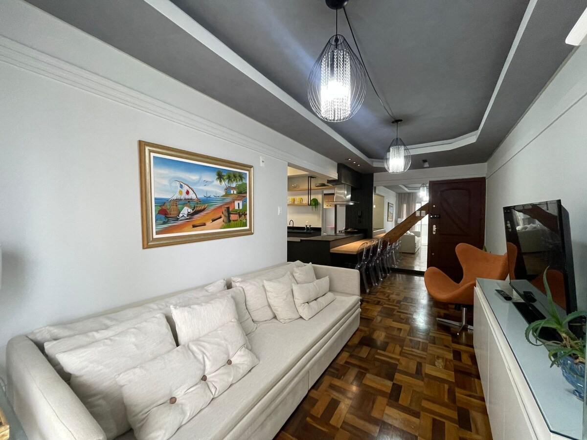 A spacious living area features a light-colored sectional sofa adorned with various-sized cushions. Natural light is offered through a nearby window, complementing the warm wood flooring. A modern kitchen is visible in the background, with a dining space that includes multiple chairs around a dark table.