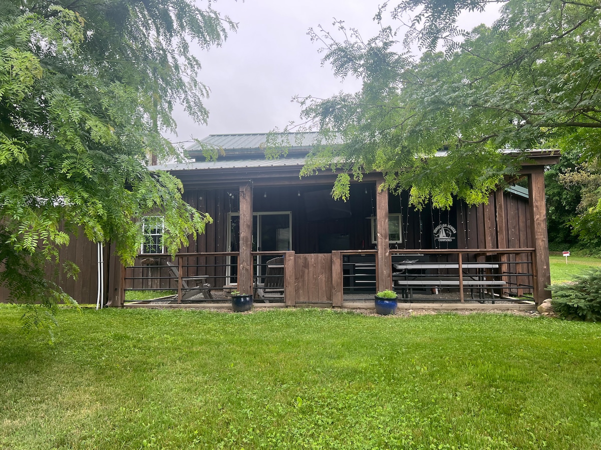 The cabin exterior is surrounded by lush greenery, with a wooden porch featuring seating arrangements. A metal roof and dark wood siding provide a rustic appearance. Potted plants are placed on the porch, adding subtle color to the outdoor space.