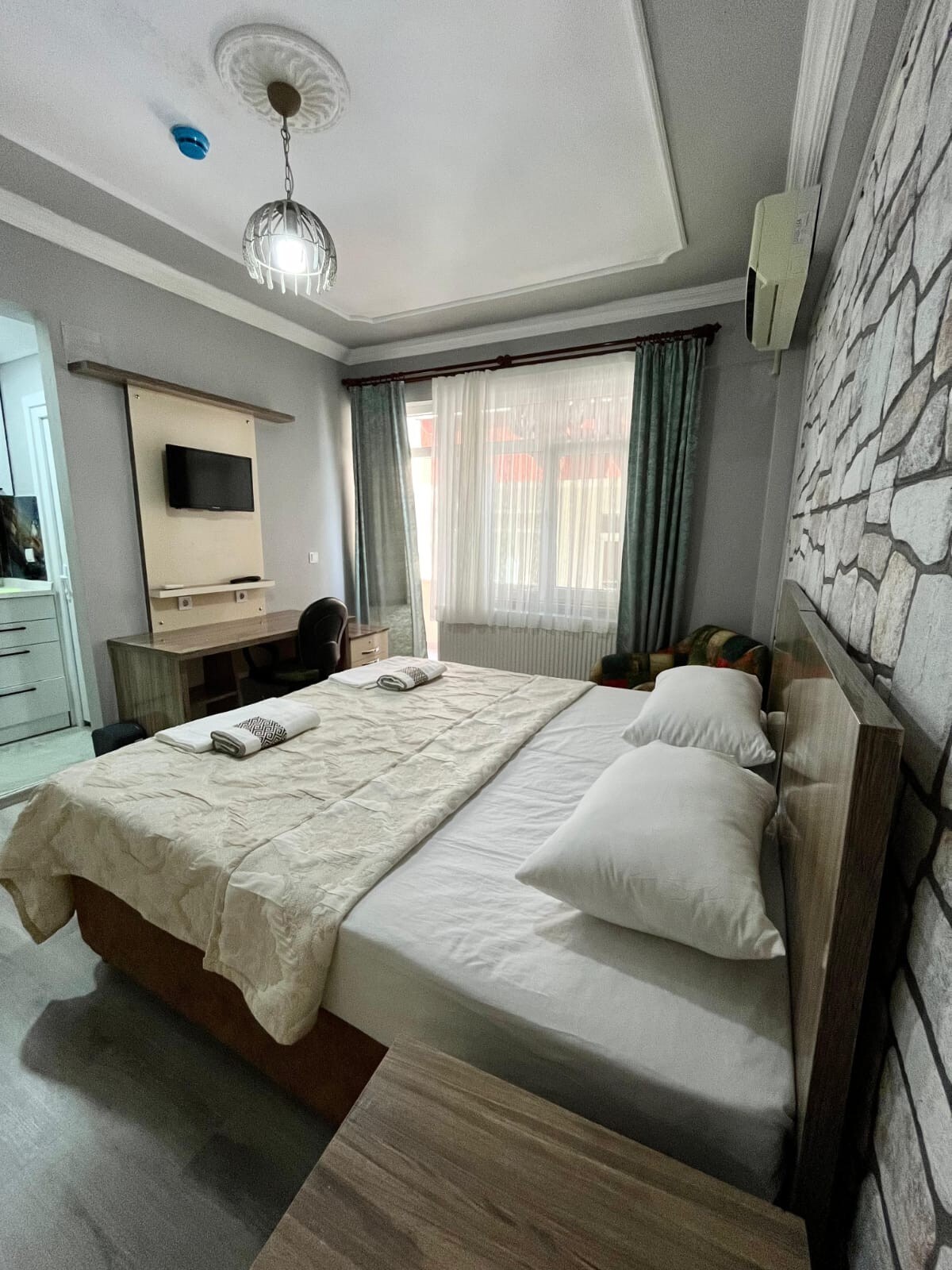 A well-lit sleeping area features a spacious bed with neatly arranged pillows and a light-colored bedspread. Two towels are placed on the bed. Natural light enters through large windows, partially covered by sheer curtains. A small desk and chair are visible in the background.