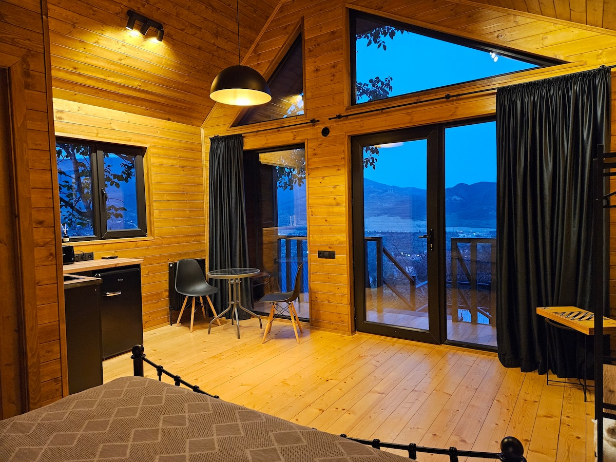 A warm wooden interior is highlighted by large windows that frame a scenic view. A dining table with two chairs is positioned near the glass doors, which open onto a balcony. Soft lighting enhances the cozy atmosphere of this stylish space.