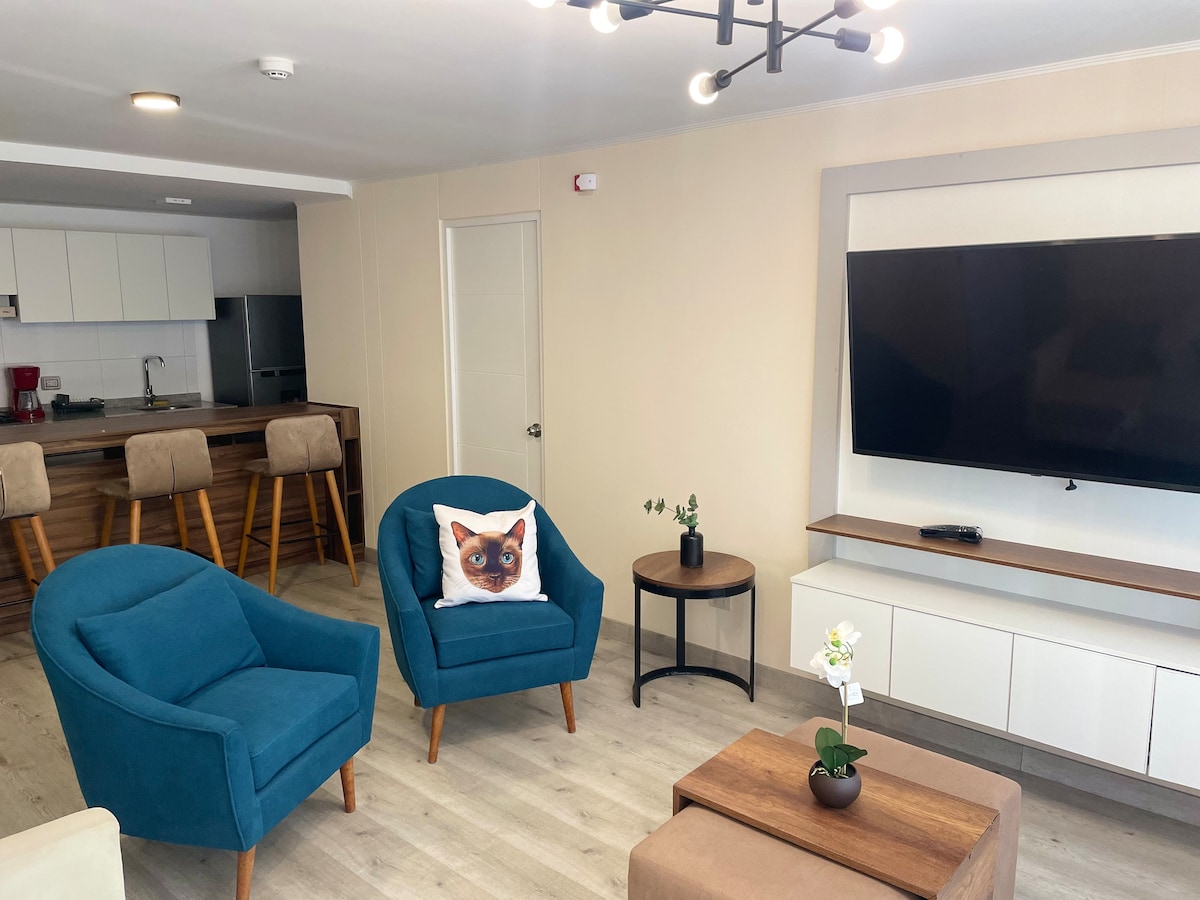 A modern living area is presented with two teal armchairs complemented by a round side table featuring a small plant. A neutral-colored sofa table supports a large, wall-mounted TV. The kitchen area is visible in the background, maintaining an open layout.