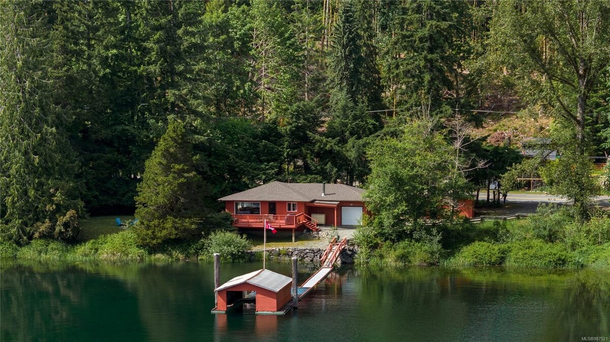 A lakefront cabin is set against a backdrop of dense green trees, with a spacious deck overlooking the water. A wooden dock extends into the lake, featuring a small boathouse. The scene is peaceful, framed by a combination of natural foliage and tranquil water reflections.
