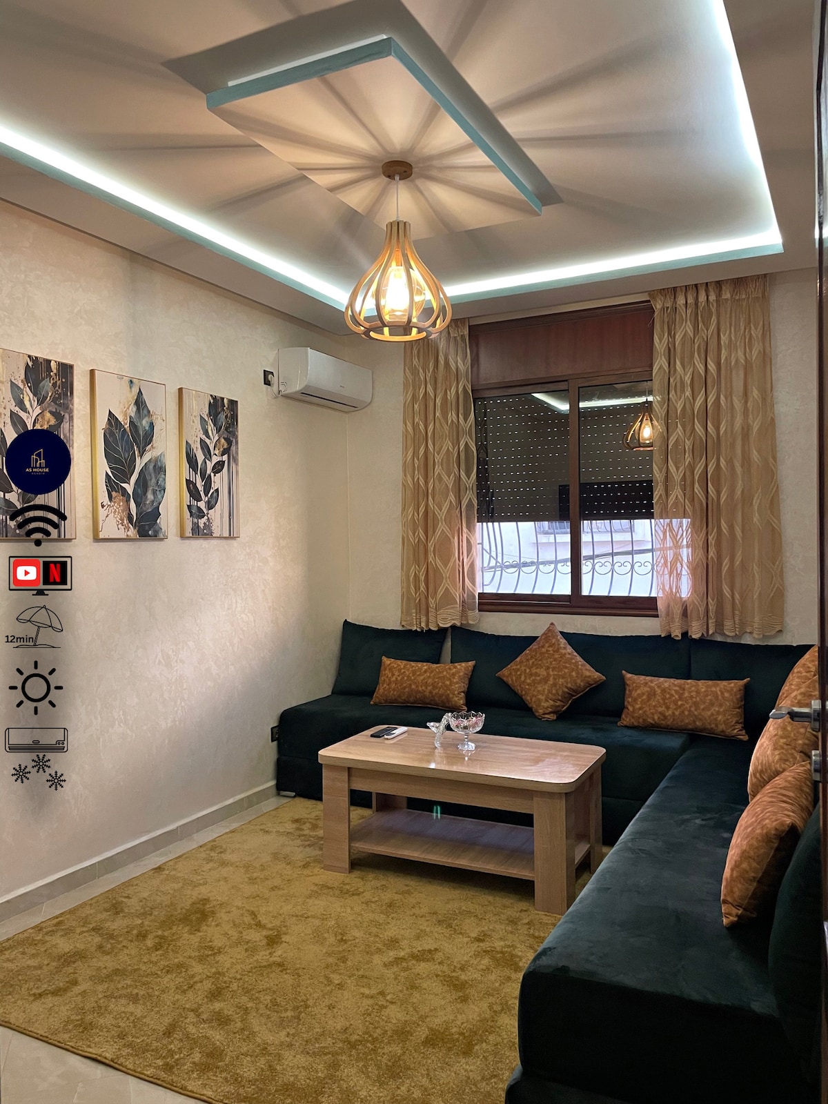 A comfortable living area features a dark green sofa adorned with patterned cushions. A wooden coffee table is positioned at the center, surrounded by a soft area rug. Artistic wall decorations and large windows with sheer curtains allow natural light to fill the space.