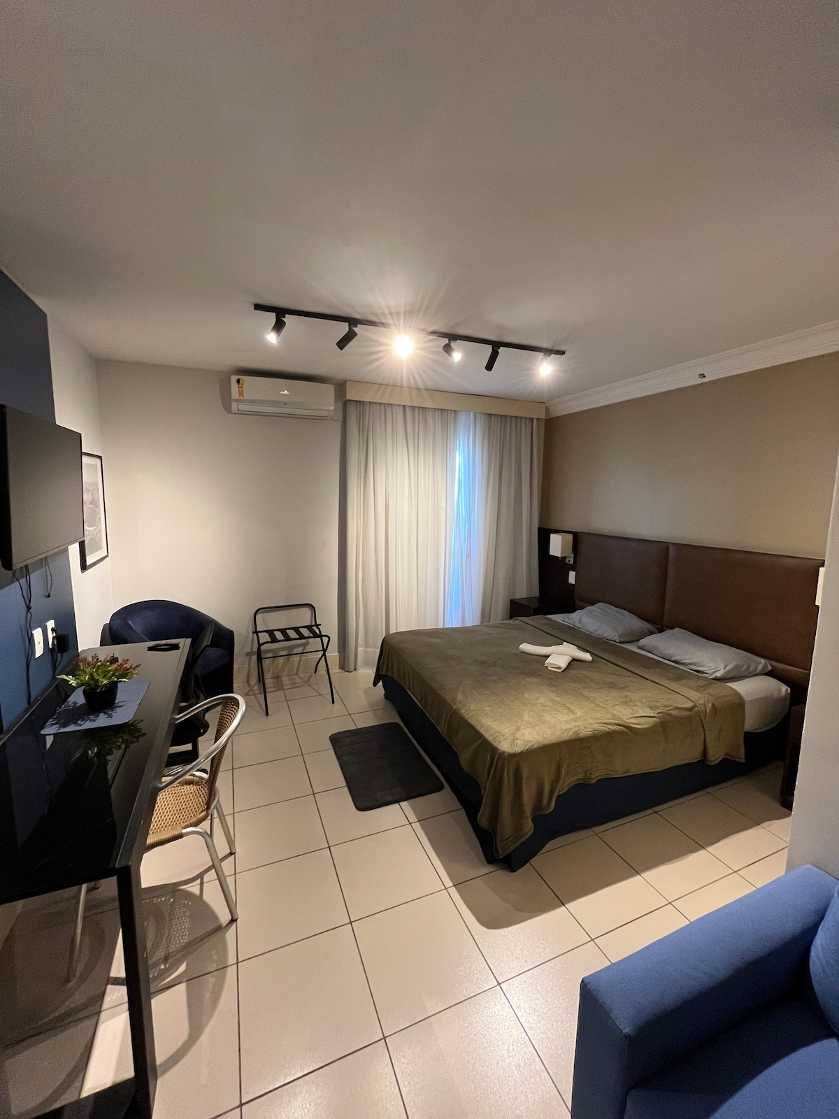 A spacious room features a comfortable queen bed with neutral bedding, complemented by a soft rug beneath. A small desk and a chair are positioned near a window, allowing natural light to filter in through sheer curtains. Air conditioning and wall-mounted television are also visible.