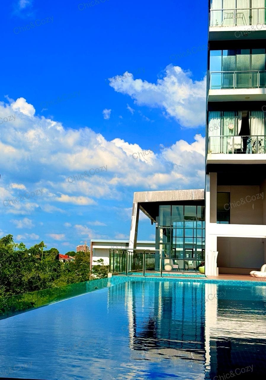 An infinity pool stretches along the edge of a modern building, reflecting the blue sky and scattered clouds above. A spacious outdoor area is visible nearby, offering a relaxing retreat with easy access to the scenic surroundings.