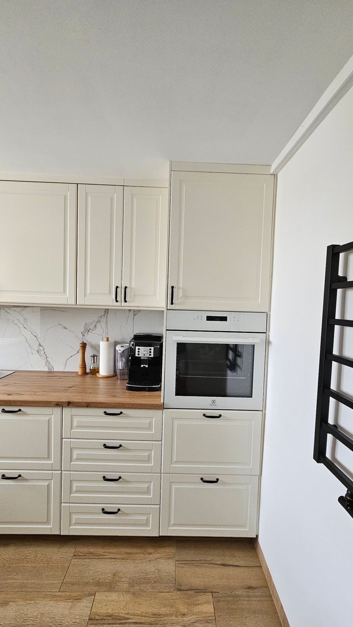 The kitchen features light-colored cabinetry with sleek black handles, complemented by a wooden countertop. Built-in appliances, including a microwave and coffee machine, are integrated into the cabinetry. A minimalist design is highlighted by a clean white wall and elegant marble backsplash.