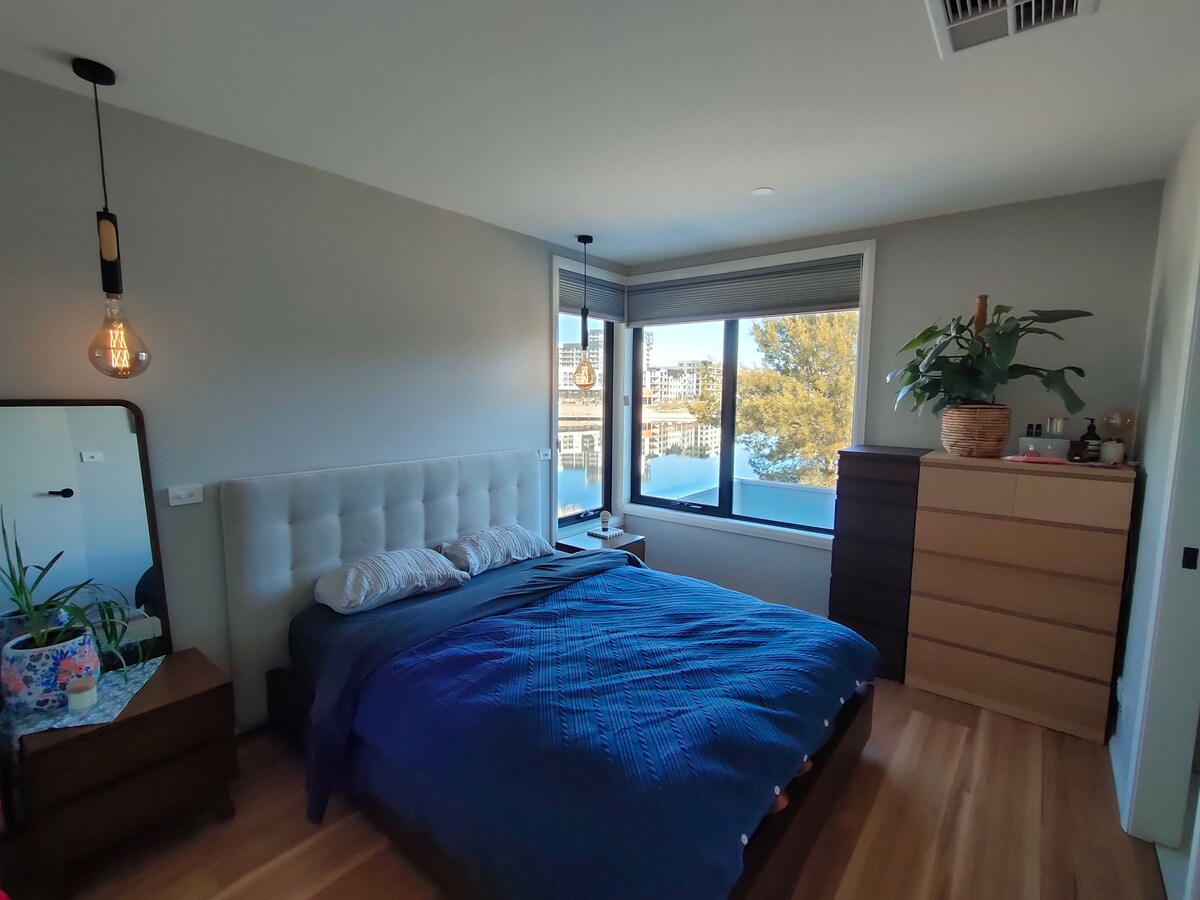 A serene bedroom is highlighted by a spacious bed dressed in blue linens, adjacent to a large window offering views of the water. A wooden dresser and a floor mirror are positioned nearby, with a potted plant adding a touch of greenery to the space.