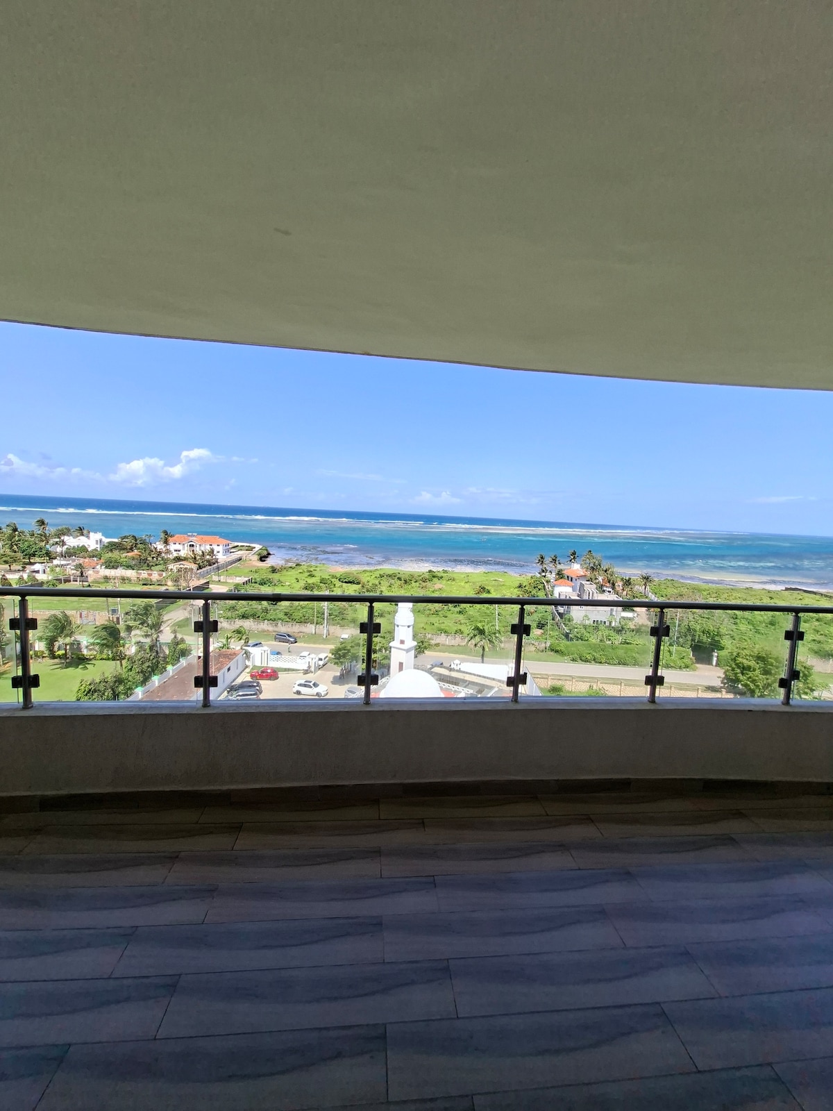 A spacious balcony offers sweeping views of the ocean and surrounding landscape. The shoreline is visible in the distance, framed by lush greenery and nearby buildings. The railing features a modern design, enhancing the open and airy feel of the space.