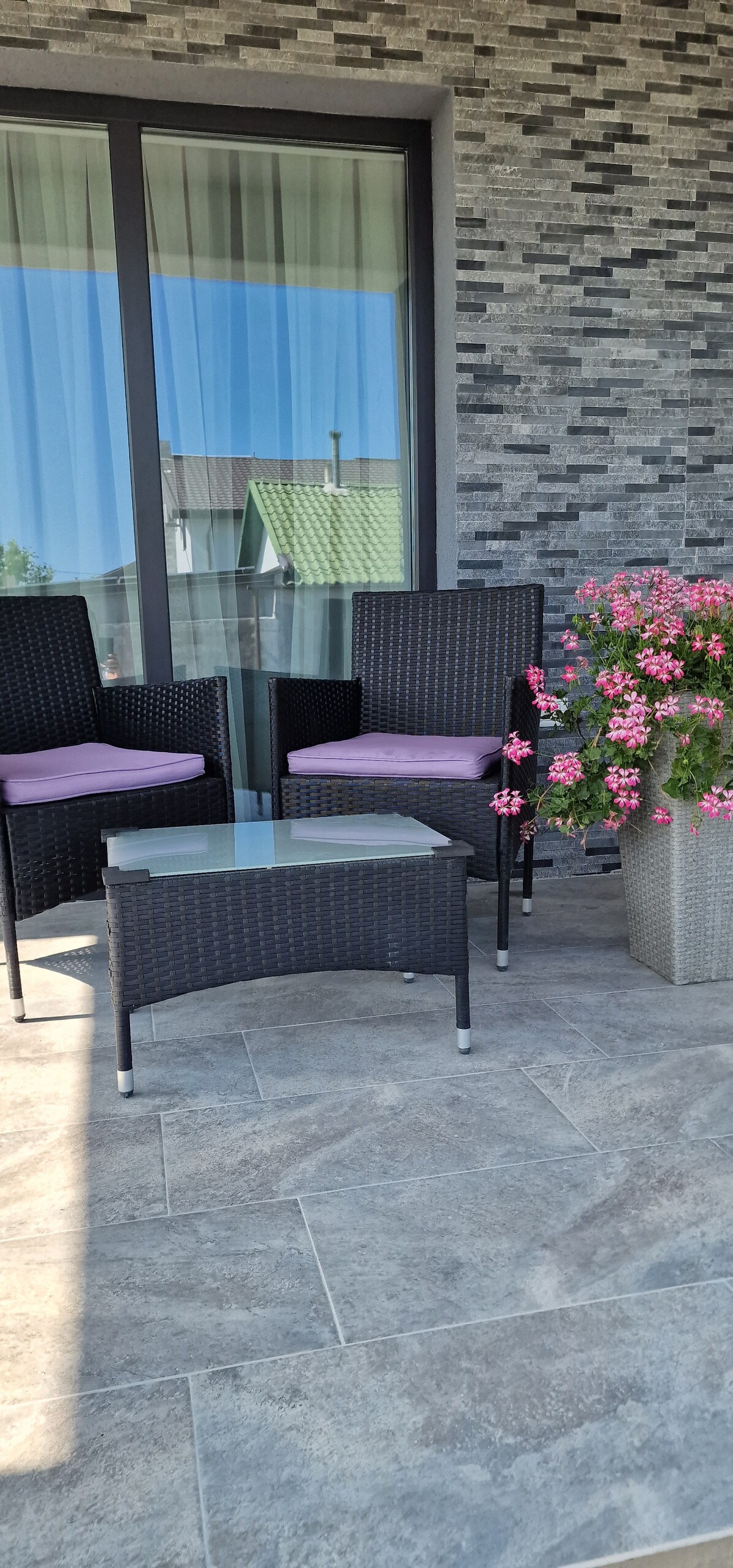 A small outdoor seating area is shown, featuring two black wicker chairs with purple cushions positioned around a glass-topped table. A flowering plant in a grey pot adds color to the scene, while a textured brick wall provides a backdrop.