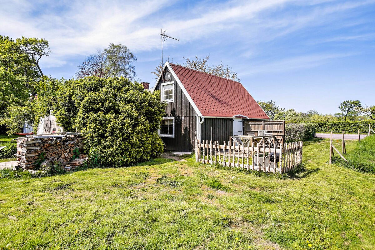 A quaint wooden cottage is set amidst lush greenery, featuring a red sloped roof. A small fenced area with a wooden gate is present, alongside a stone feature. The surrounding landscape is open and includes trees and grass, enhancing the natural setting.