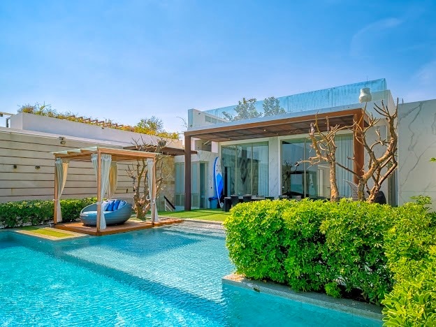 An inviting outdoor space features a swimming pool surrounded by lush greenery. Sleek lounge chairs are positioned under a wooden pergola, while a clear sky complements the modern architectural design of the nearby villa. Glass walls provide a view into the interior living area.