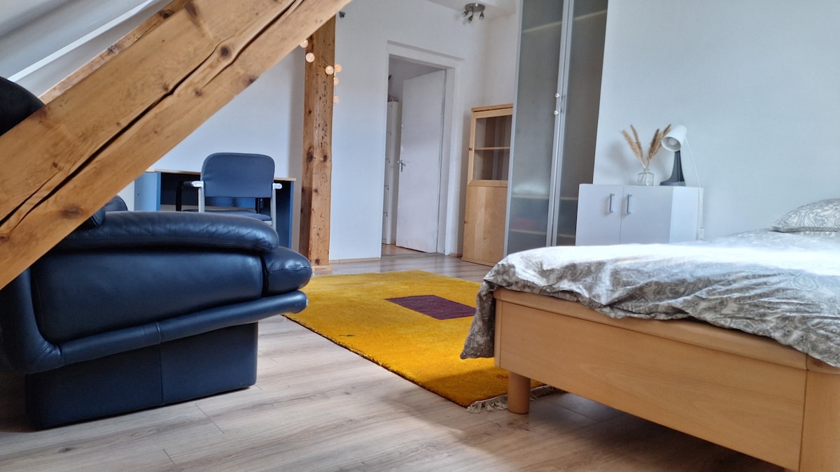 A spacious living area features a large bed with a light duvet, a dark leather sofa, and a wooden desk. A textured rug in yellow and brown adds warmth. Light enters through a nearby window, with wooden beams contributing to the room's character.