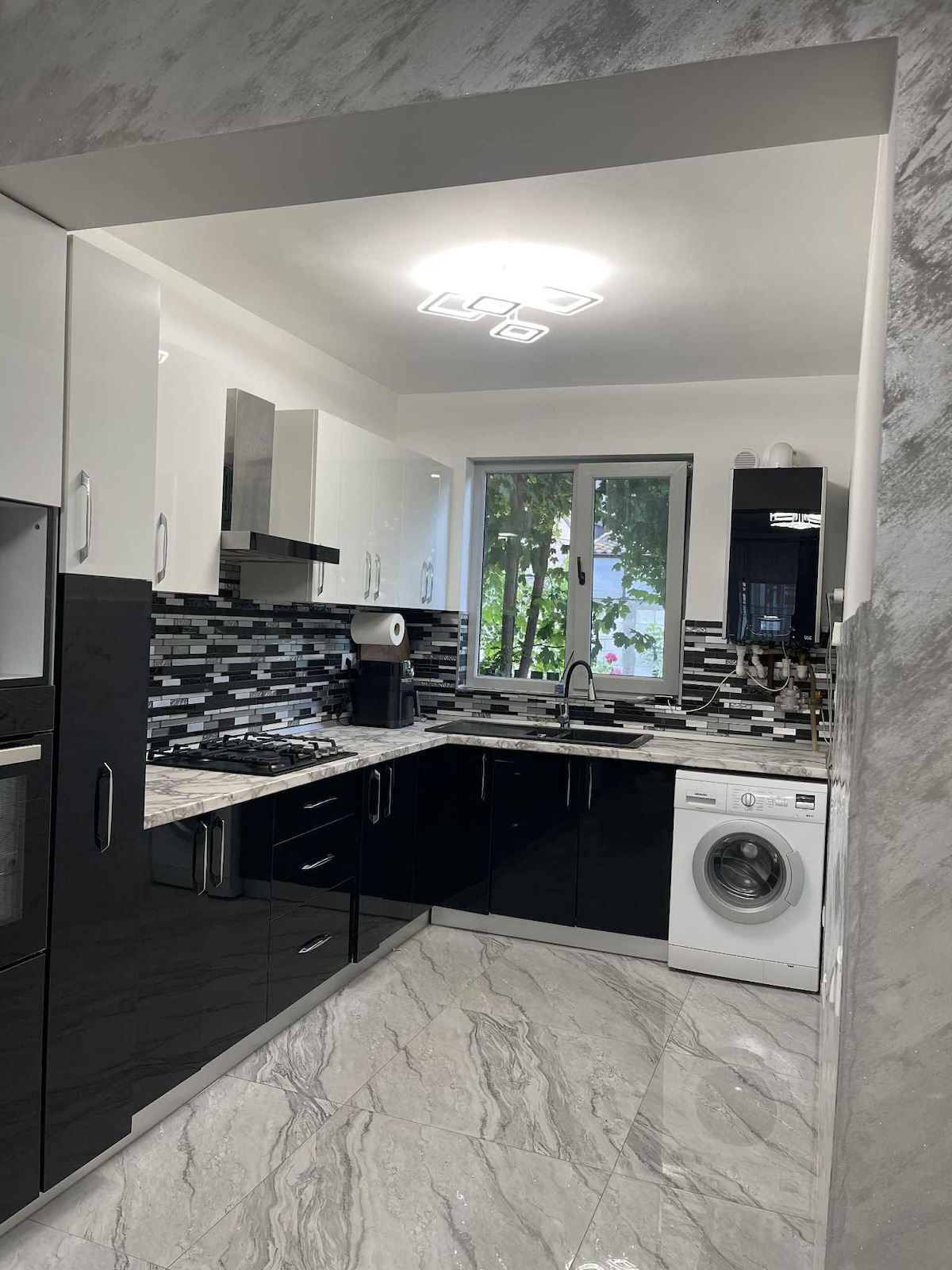 A modern kitchen features sleek black and white cabinetry with a marble countertop. Stainless steel appliances are installed, including a gas stove and an oven. A washing machine is positioned near the window, which provides natural light and views of greenery outside.