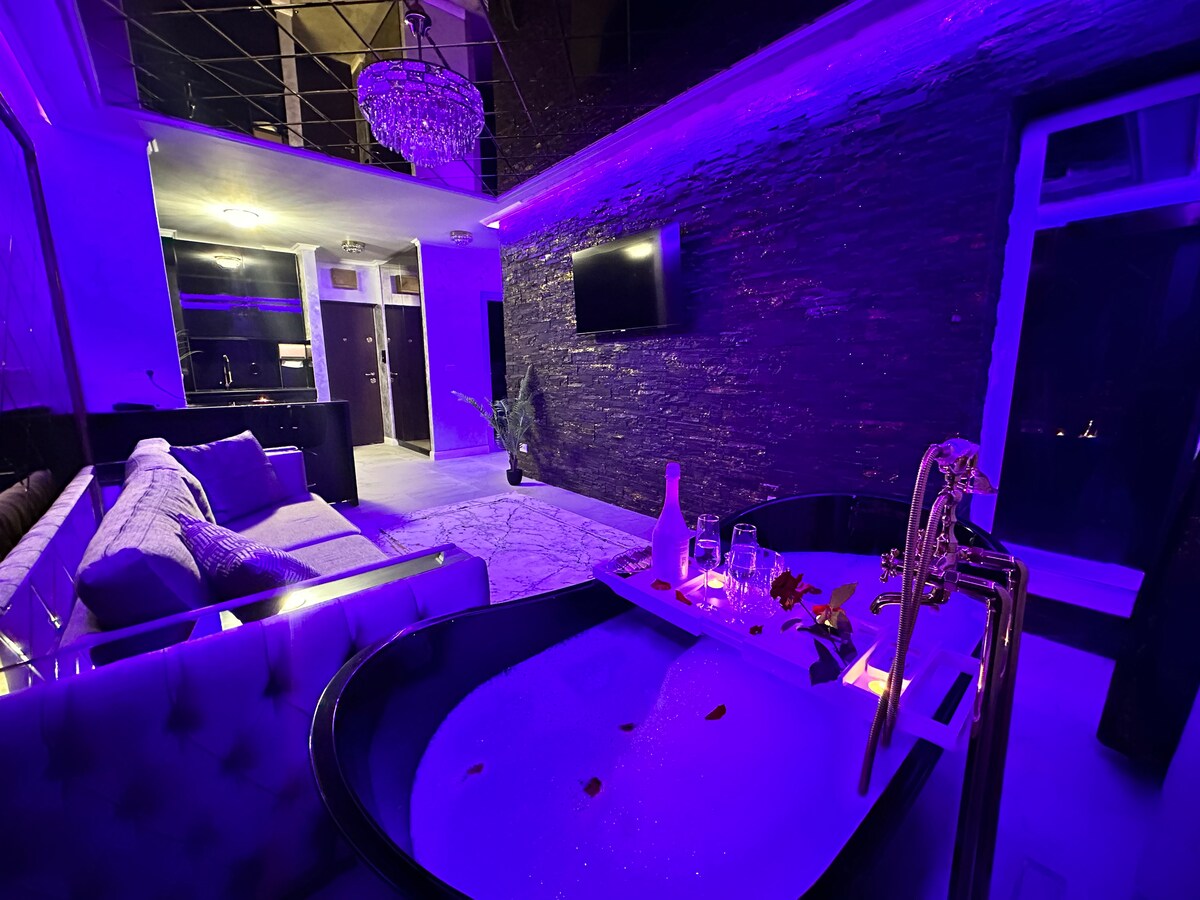 An inviting space features a freestanding bathtub adorned with rose petals, illuminated by adjustable LED lights. A cozy seating area with a sofa is complemented by a modern television mounted on a textured wall. Kitchen amenities are visible in the background.