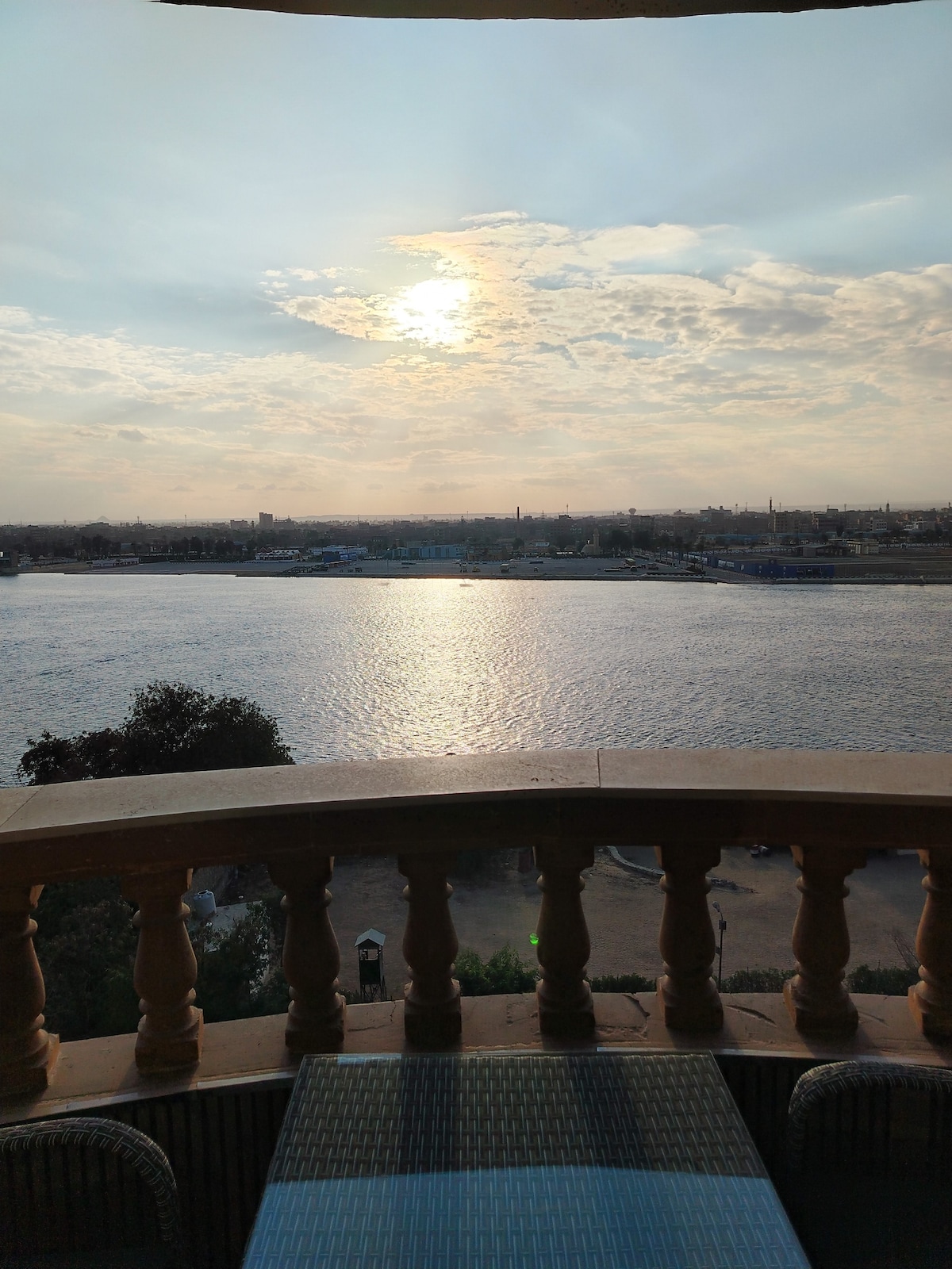 A scenic view over the Nile River is captured from a balcony, showcasing a soft evening light reflecting on the water. The horizon is dotted with a blend of natural and urban landscapes under a sky with scattered clouds.