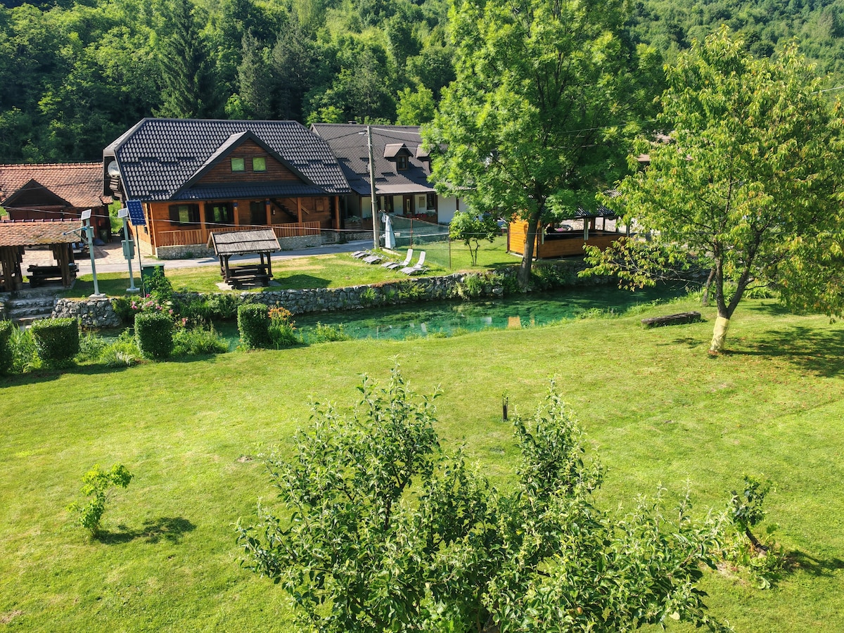 A scenic view captures a well-maintained grassy area with trees and a serene river running through it. Several wooden structures are visible, including a large house with a patio and seating, and a shaded area featuring chairs for relaxation.