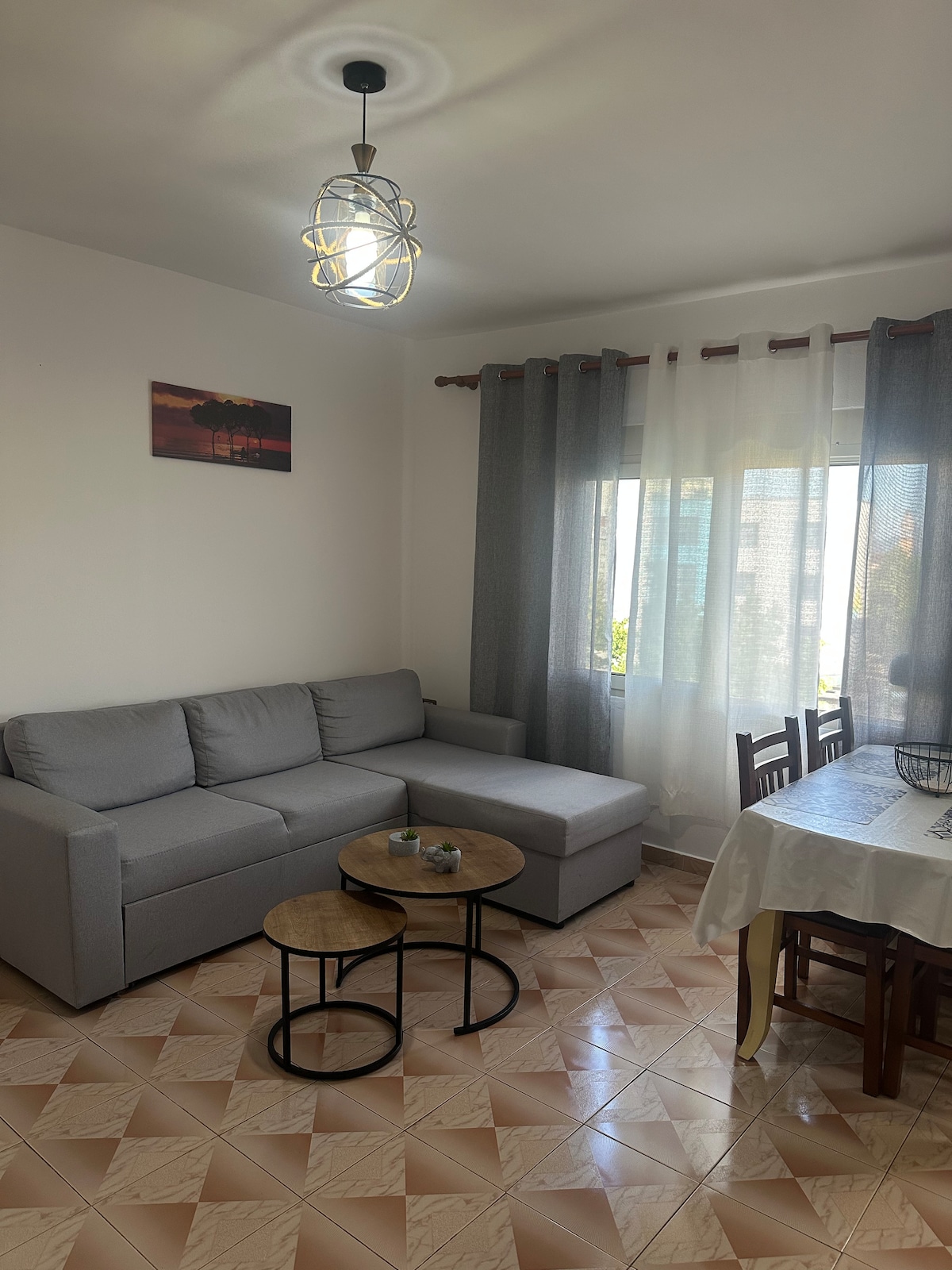 A cozy living area features a gray sectional sofa positioned next to a window with sheer curtains, allowing natural light to filter in. Two round tables are placed in front of the sofa, while a dining table with wooden chairs is visible in the corner.