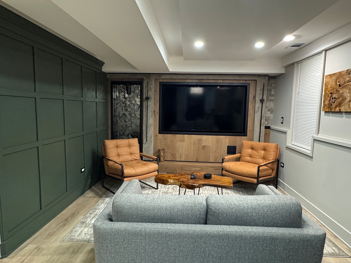 A modern living area features a large flat-screen TV mounted on a wooden wall. Two tan armchairs sit opposite a gray sofa, creating a comfortable seating arrangement. A wooden coffee table occupies the center, and textured walls in green add depth to the space.