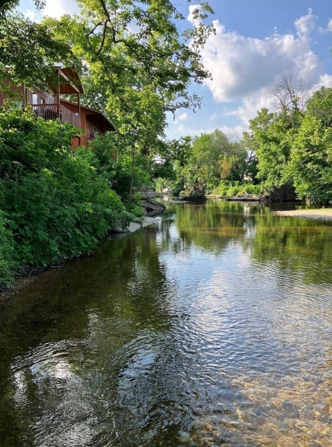 A serene view of a calm river surrounded by lush greenery and trees. The clear water reflects the sky and foliage, creating a peaceful atmosphere. A wooden structure is visible in the background, blending harmoniously with the nature surrounding it.