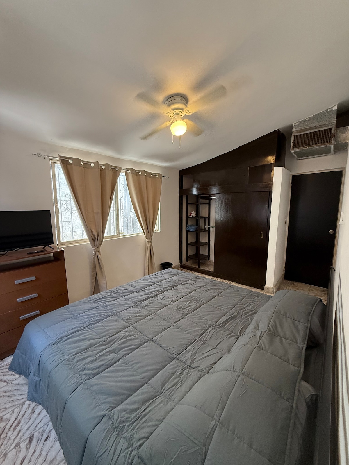 A comfortable bedroom features a spacious bed covered with a grey blanket. Natural light enters through two windows adorned with light drapes. A dresser and TV are positioned against the wall, while a shelf stands nearby, providing additional storage options.