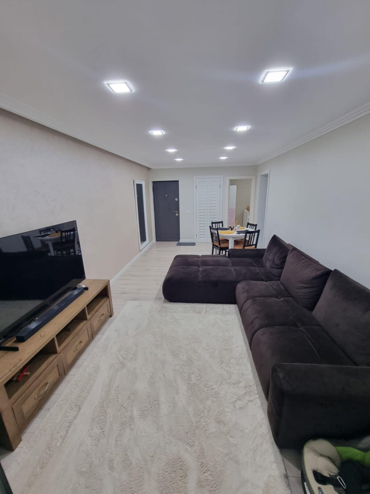 A spacious living area is depicted, featuring a dark sectional sofa positioned alongside a wooden TV stand with a flat-screen television. A dining area with a table and four chairs is visible in the background, complemented by neutral-colored walls and soft lighting.