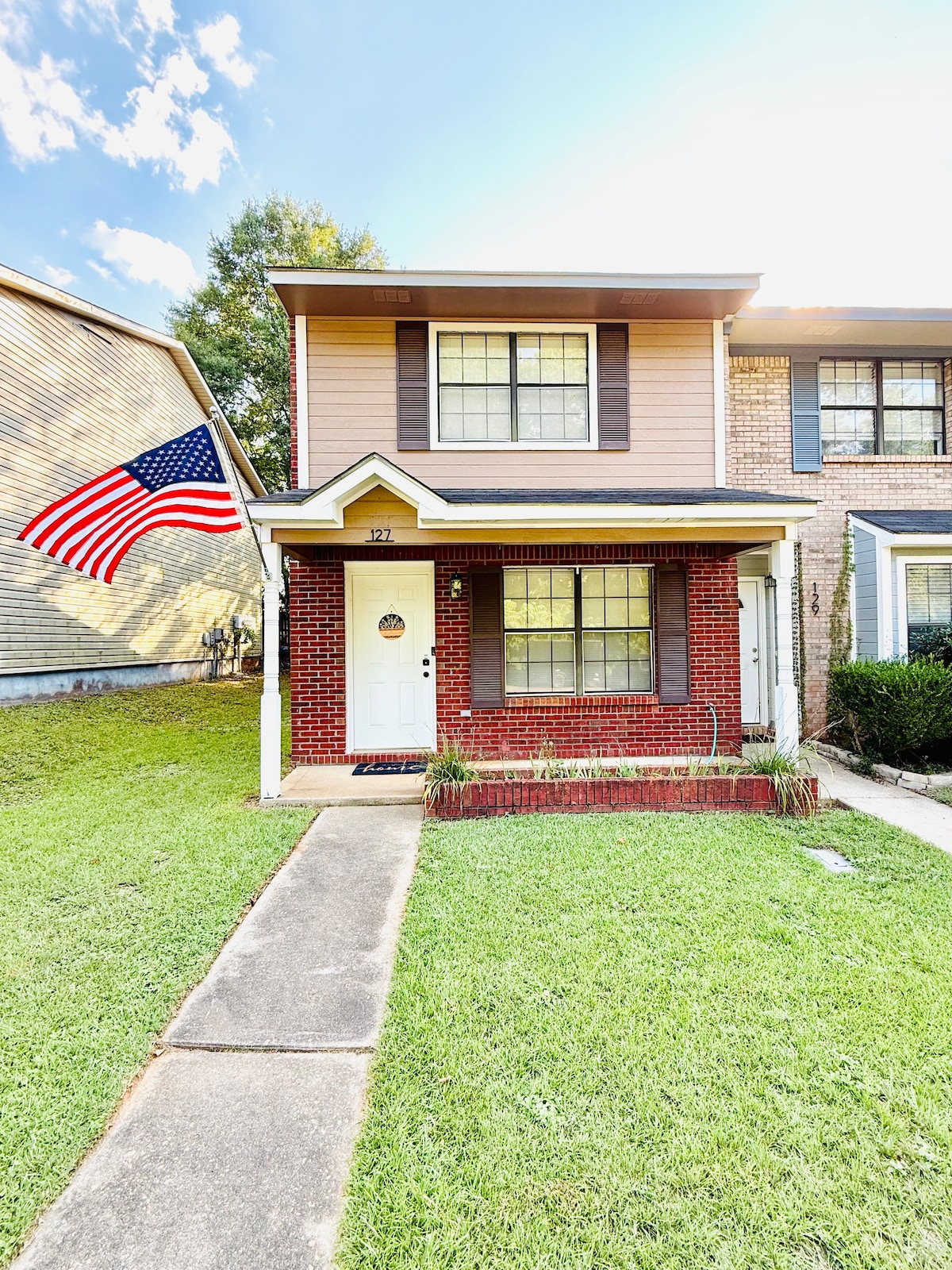 The exterior of a two-story home is shown, featuring a welcoming entryway with a white door and multiple windows. A large American flag flutters from the front porch. The green lawn is well-maintained, with a concrete pathway leading to the entrance.