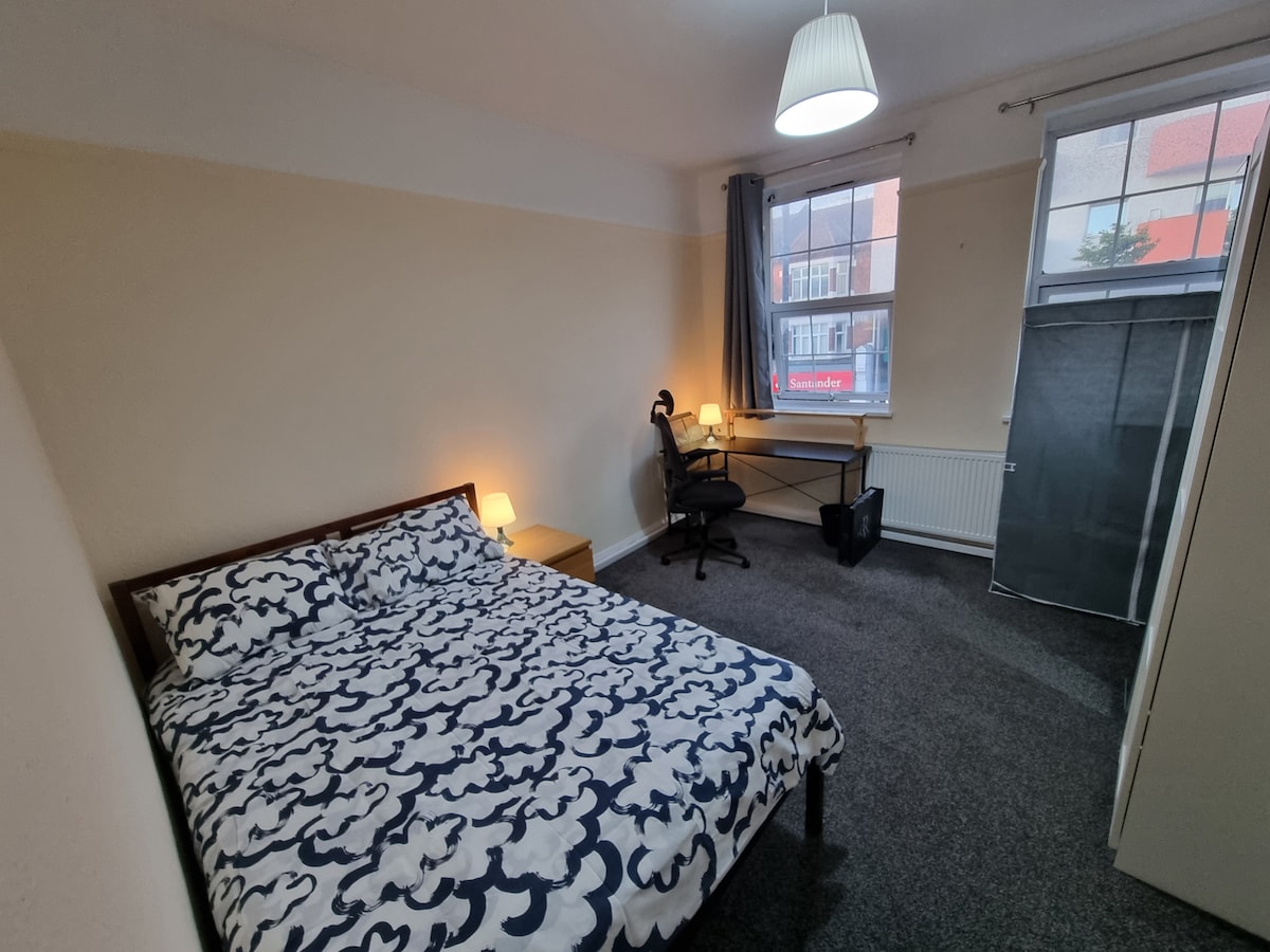 A spacious private room features a double bed with a patterned duvet cover. A desk with a chair is positioned by the window, allowing natural light to fill the space. Two bedside lamps provide additional lighting, enhancing the overall comfort of the room.