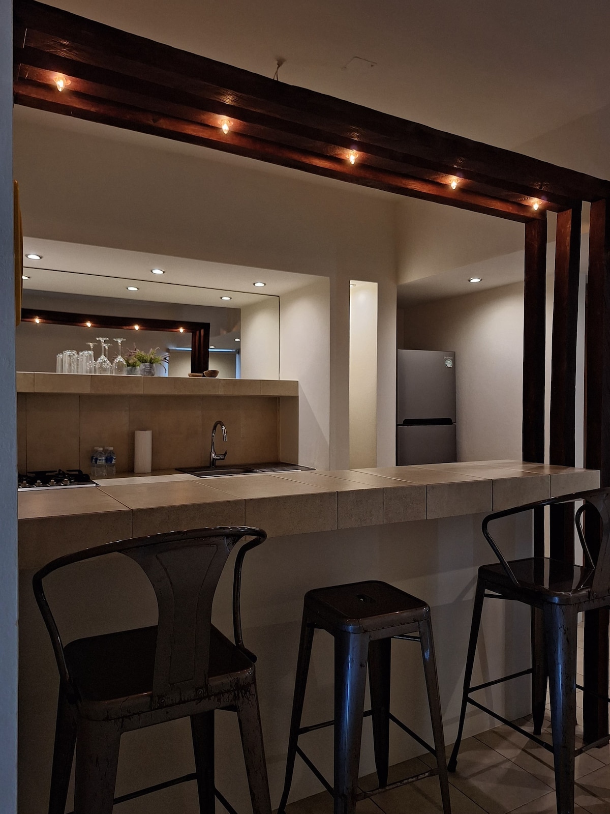 An open kitchen area features a bar counter with three high stools, illuminated by subtle ceiling lights. A modern refrigerator is visible in the background, while kitchen utensils are neatly arranged on the counter.