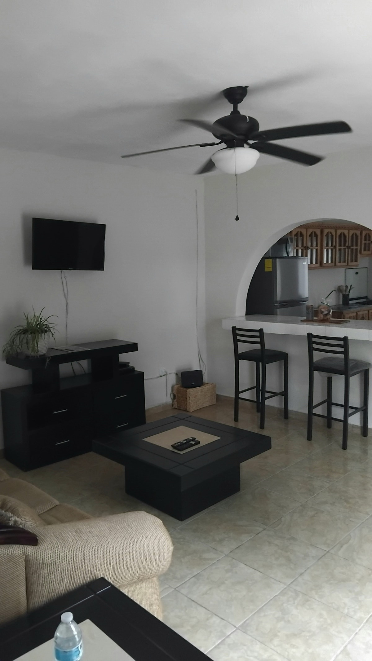 A living area showcases a light-toned sofa oriented towards a wall-mounted television. Nearby, a black coffee table is positioned on tiled flooring. A small plant adds a touch of greenery. Behind, high-top bar stools are arranged at a kitchen counter, enhancing the open layout.