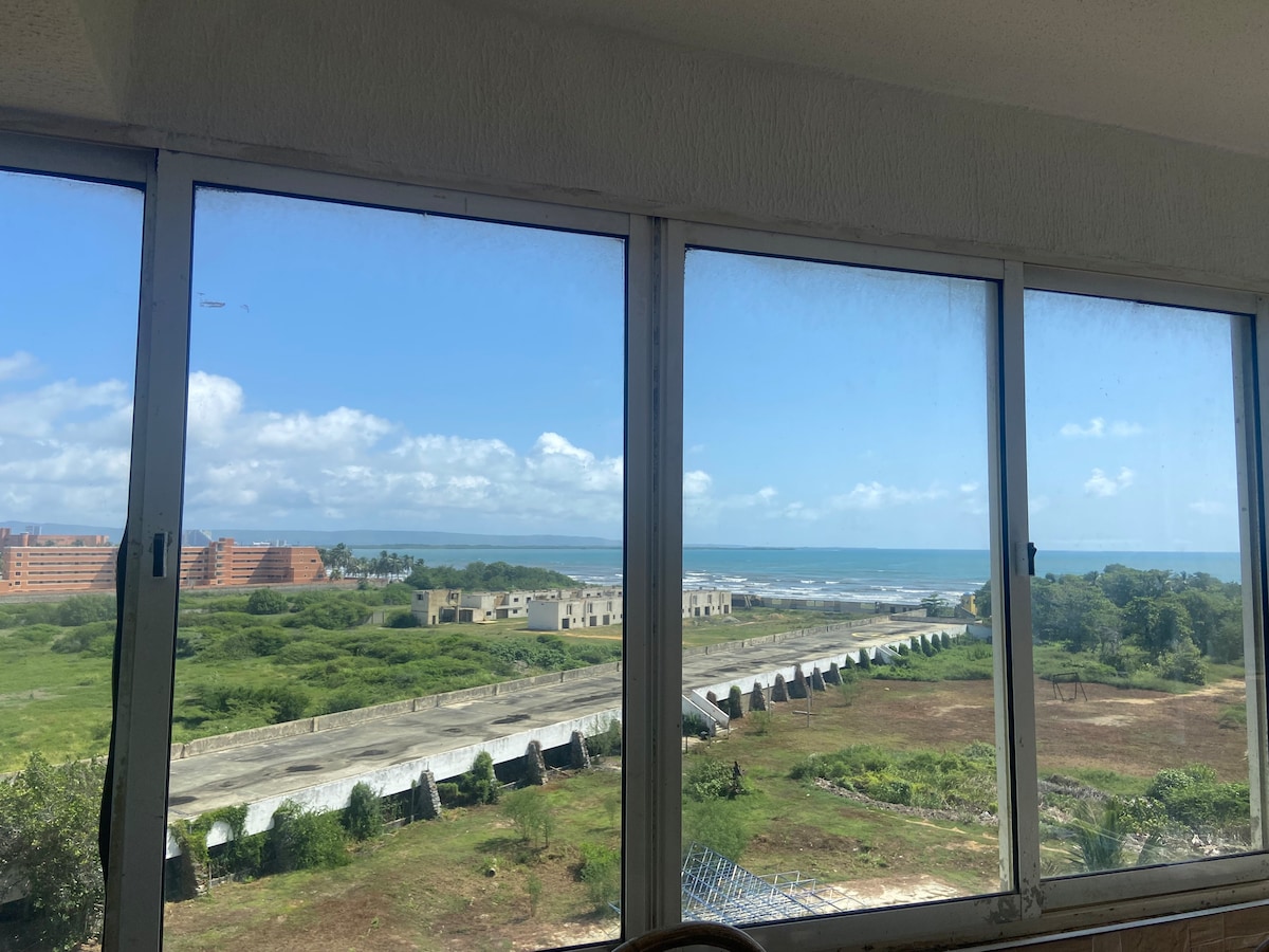A large window provides a panoramic view of the ocean and surrounding landscape. The skyline features a mix of green spaces and low-rise buildings, with the beach visible in the distance. Bright blue skies and scattered clouds enhance the outdoor scene.