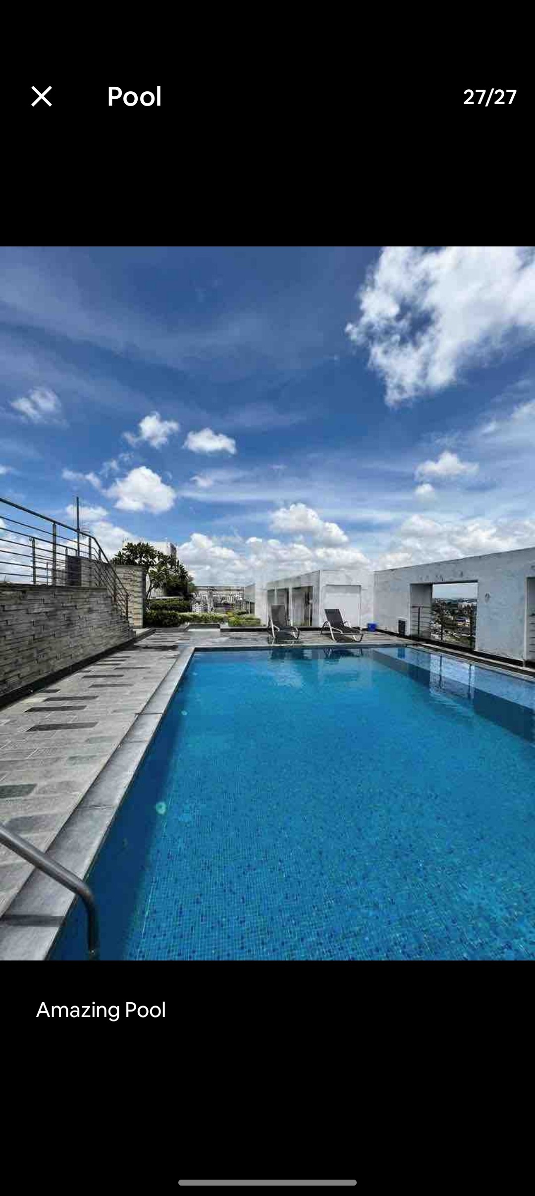 A tranquil pool area is highlighted with shimmering blue water, surrounded by sleek stone flooring. Comfortable loungers are positioned nearby, and lush greenery can be seen in the background. The open sky adds a bright, airy feel to the space.