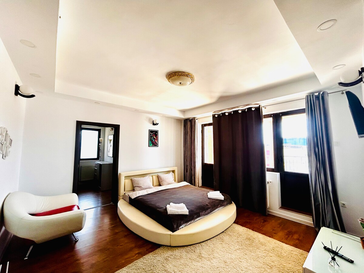 A spacious bedroom features a round bed with a dark comforter and two folded towels on top. Large windows allow natural light to fill the room, adorned with dark curtains. A light-colored rug covers the floor, and a modern chair is positioned nearby.