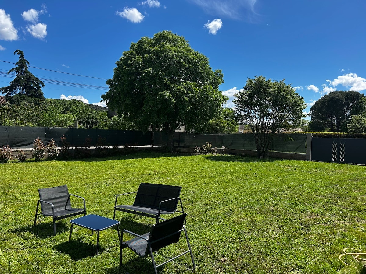 A spacious private garden is featured, with a lush green lawn and a clear blue sky overhead. Two black chairs and a small table are arranged on the grass, inviting relaxation. Trees and shrubs are visible along the garden's perimeter, offering a sense of tranquility.