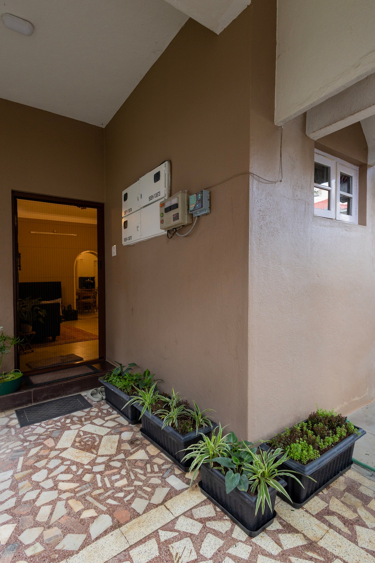 A welcoming entrance features a brown exterior wall and a neatly arranged line of potted plants along a patterned stone pathway. Nearby, a door opens to reveal the interior of the home, with mailboxes mounted on the wall for convenience.