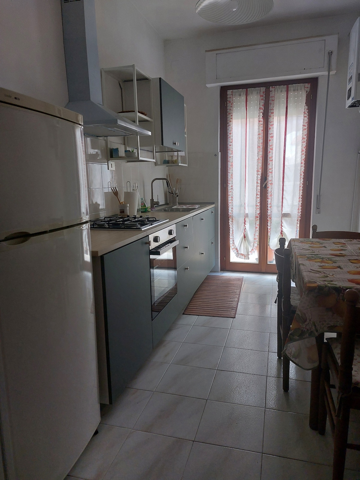 A compact kitchen features modern cabinetry and a gas stove. A refrigerator stands alongside a countertop with essential appliances. Natural light enters through a window, complemented by a balcony door adorned with patterned curtains. A wooden dining table occupies the space, surrounded by several chairs.