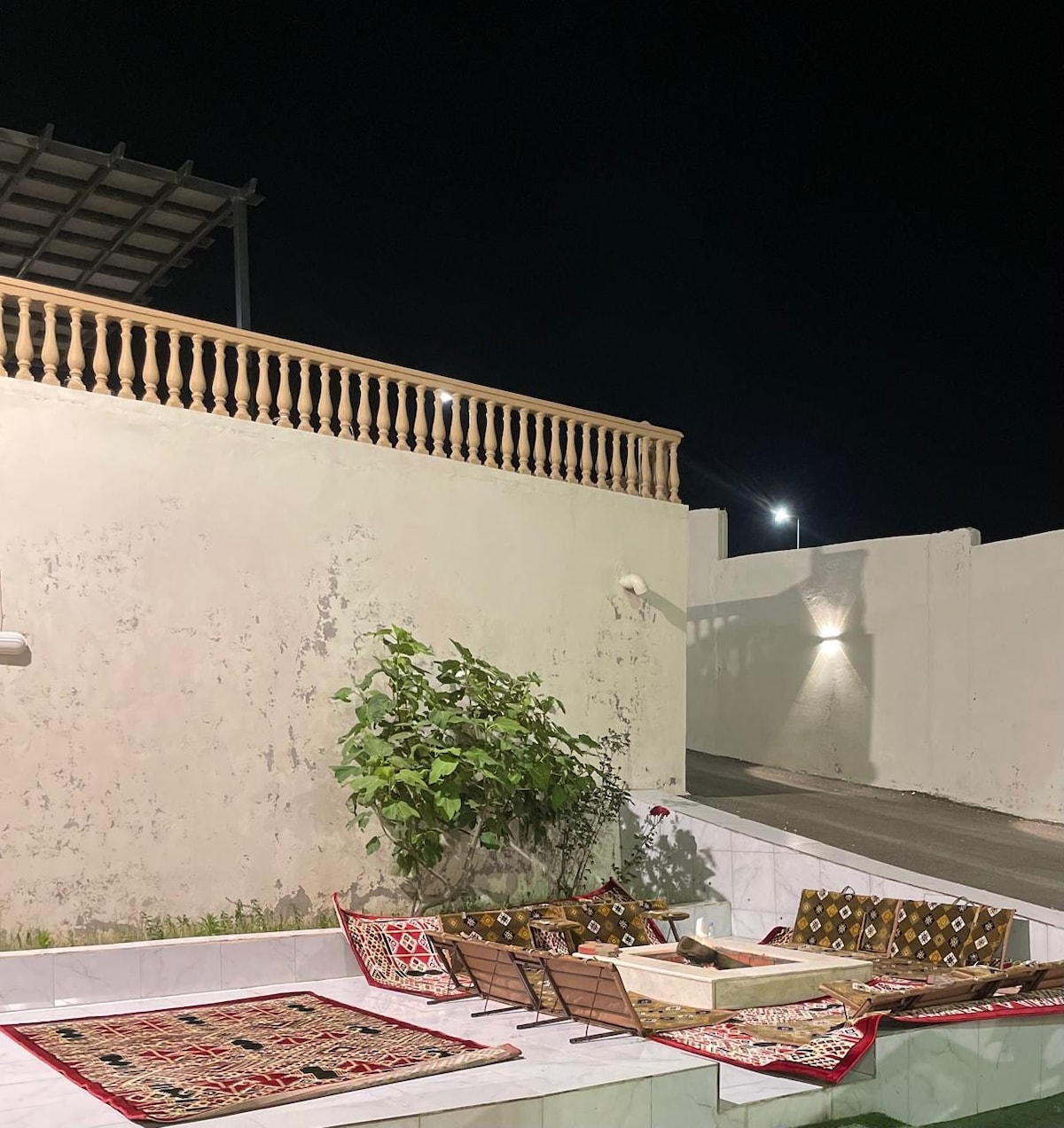 An outdoor seating area features a series of low benches adorned with traditional fabrics. The walls are painted in a light hue, complemented by a green plant. Soft lighting illuminates the area, creating a serene atmosphere at night.