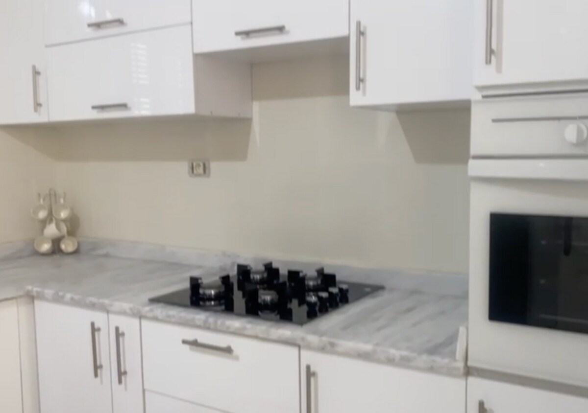 A modern kitchen showcases sleek white cabinetry with polished silver handles. A gray marble countertop features a black gas stovetop with multiple burners. An oven built into the cabinetry is visible, adding to the functional design of the space.