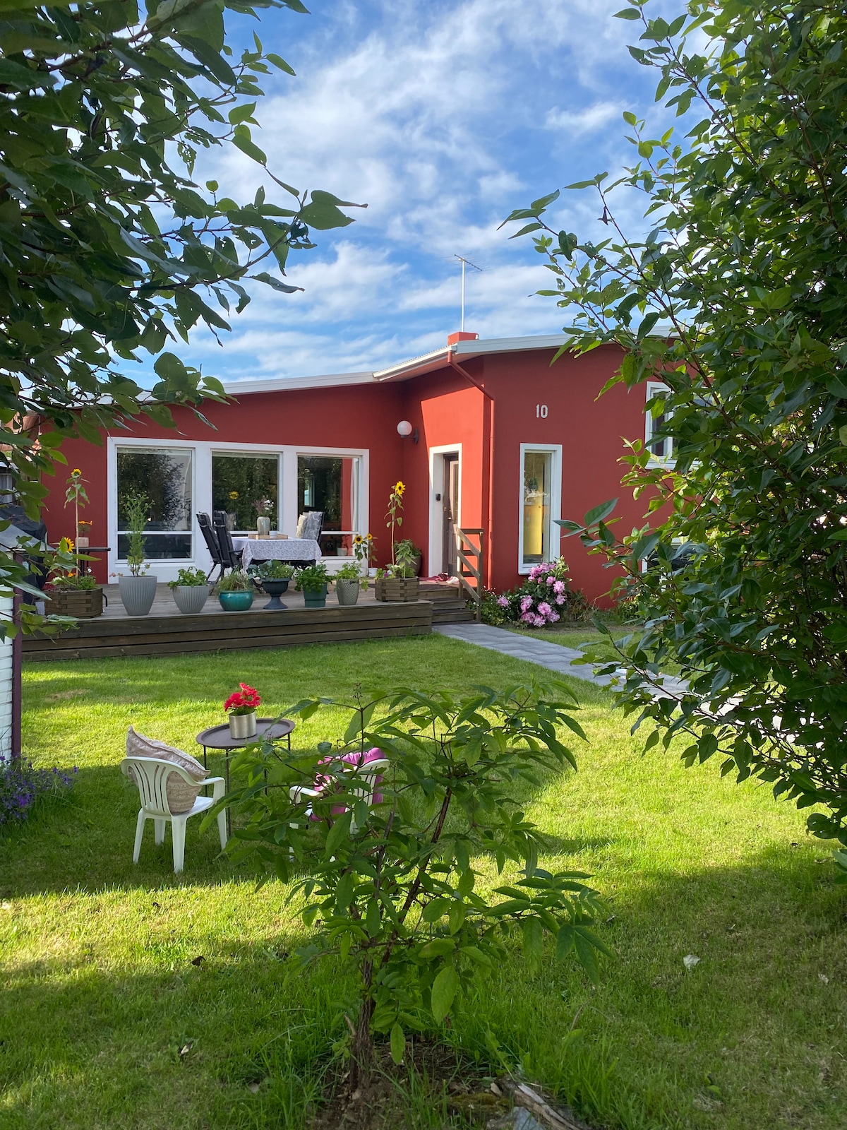 A vibrant red home is framed by lush greenery and colorful flowerbeds. A spacious outdoor deck features seating areas and potted plants. A well-maintained lawn provides ample space for outdoor activities, while a clear blue sky adds a bright backdrop to the inviting scene.