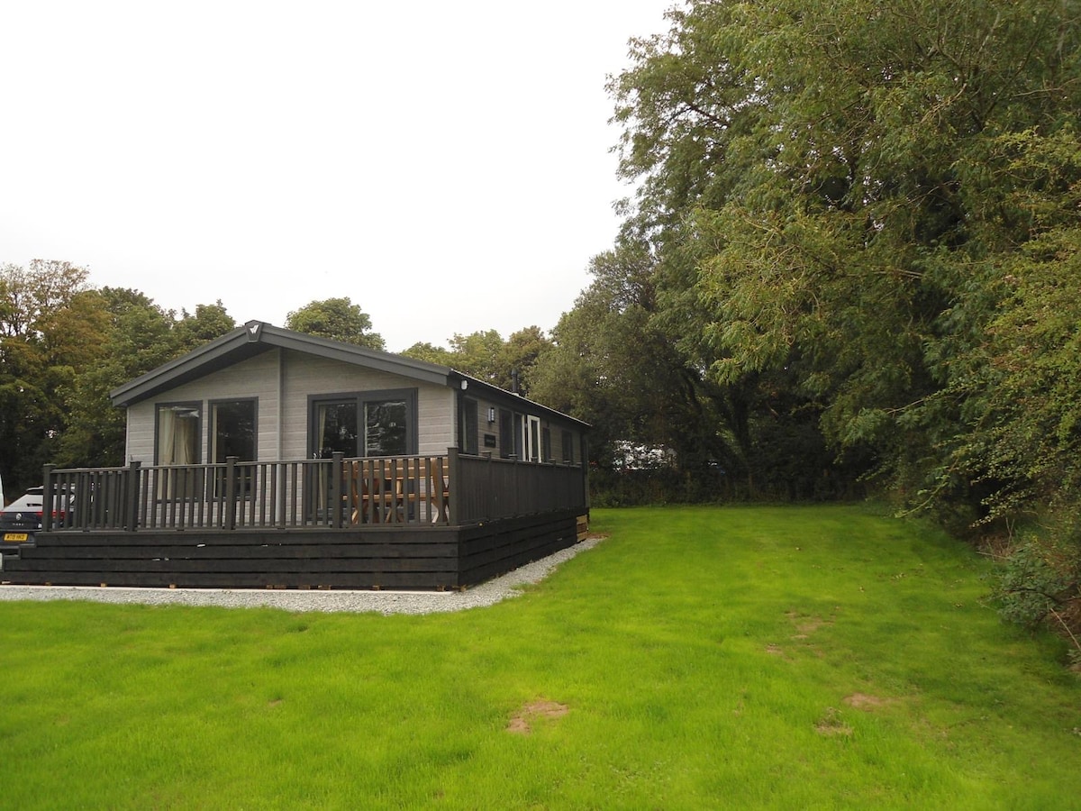 A charming holiday lodge is nestled amidst greenery, featuring a spacious wooden deck that wraps around its exterior. Lush grass surrounds the lodge, and trees provide a natural backdrop, enhancing the serene countryside setting.