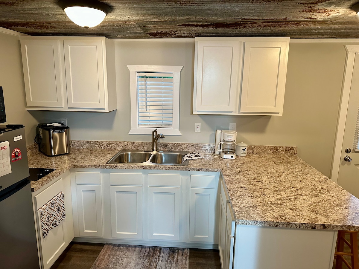 A bright kitchen space features white cabinetry with modern design and a large countertop. Stainless steel appliances include a refrigerator and a coffee maker. A double sink is positioned beneath a window, allowing natural light to illuminate the area, enhancing the welcoming environment.