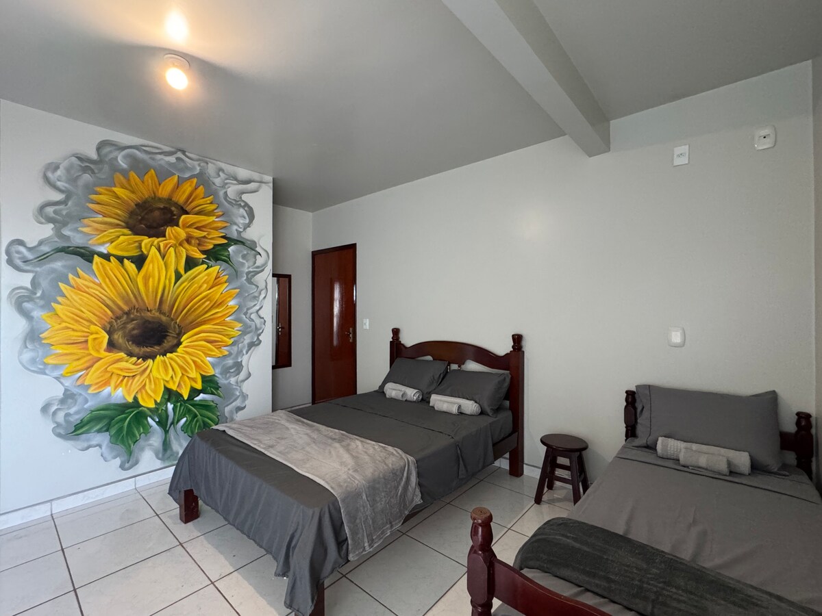 A spacious sleeping area features a large double bed and a smaller single bed, both dressed in neutral linens. A mural of vibrant sunflowers adds a touch of color to the otherwise neutral walls. A small wooden stool is positioned beside the beds.